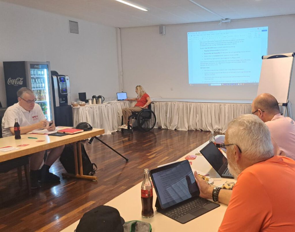 Ein Konferenzraum mit mehreren Teilnehmenden einer Sitzung. Im Vordergrund sitzen drei Männer an einem Tisch und arbeiten konzentriert an Laptops und Tablets. Zwei von ihnen tragen orangefarbene Oberteile. Auf dem Tisch stehen Getränkeflaschen, Gläser und Snacks.
Im Hintergrund, auf der linken Seite, sitzt ein Mann im weißen SPD-Poloshirt mit einer Cola-Flasche vor sich. Auf der rechten Seite des Raums sitzt eine Frau im Rollstuhl an einem separaten Tisch mit Laptop. Hinter ihr ist eine weiße Tischreihe mit Getränken, Kaffeekannen und Gläsern aufgebaut.
An der Wand wird ein Textdokument per Beamer projiziert – offenbar ein Sitzungspunkt oder eine gemeinsame Arbeitsvorlage. Die Atmosphäre wirkt ruhig, konzentriert und gut strukturiert.