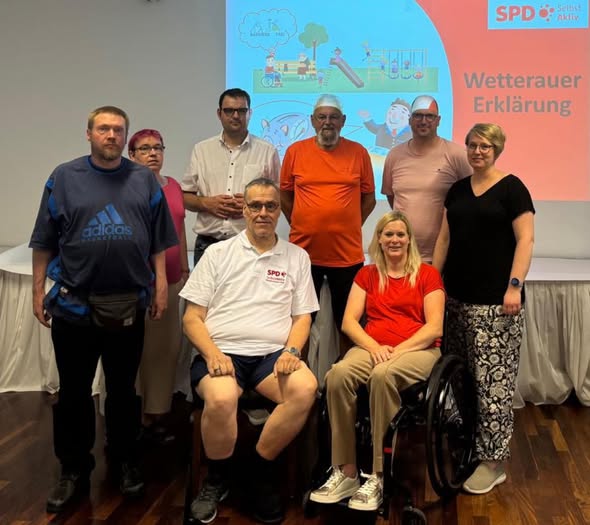 Vorstand der Selbst Aktiv Hessen-Süd gemeinsam in einem Veranstaltungsraum. Im Hintergrund ist eine große Präsentationsfolie auf eine Leinwand projiziert. Oben rechts auf der Folie steht das Logo der SPD Selbst Aktiv sowie der Titel „Wetterauer Erklärung“. Links auf der Folie sind bunte Illustrationen von Menschen mit und ohne Behinderung auf einem Spielplatz zu sehen.
Die Gruppe besteht aus sieben stehenden und zwei sitzenden Personen. Eine der sitzenden Personen ist eine Frau im Rollstuhl. Alle blicken in die Kamera und lächeln oder schauen freundlich. Die Kleidung ist sommerlich und locker, die Stimmung wirkt konzentriert und positiv.