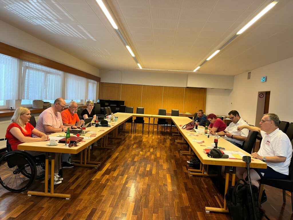 Neun Personen sitzen einander gegenüber an zwei langen Tischen in einem Tagungsraum. Sie wirken konzentriert und arbeiten mit Laptops, Tablets und Notizen. Die linke Tischseite ist überwiegend weiblich besetzt, die rechte eher männlich. Am Tischende rechts sitzt ein Mann im weißen SPD-Poloshirt mit Gehhilfe neben sich. Links im Bild sitzt eine Frau im Rollstuhl.
Auf den Tischen stehen Getränke, Tassen und Arbeitsmaterialien mit SPD-Logo. Der Raum ist hell, mit großen Fenstern und Holzfußboden. Die Atmosphäre wirkt ruhig und produktiv – ein Moment aus der Klausurtagung der SPD AG Selbst Aktiv im Bezirk Hessen-Süd.