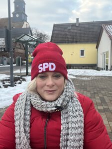 Ich mit Roter SPD Mütze, Schal und Dicker Jacke auf einem Vorplatz. Hinter mir Gebäude Bushaltestelle, Straße und eine Kirche. Es liegt vereinzelnd Schnee